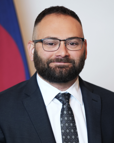 Tony Gherardini wearing a black suit and smiling in front of a Colorado flag.